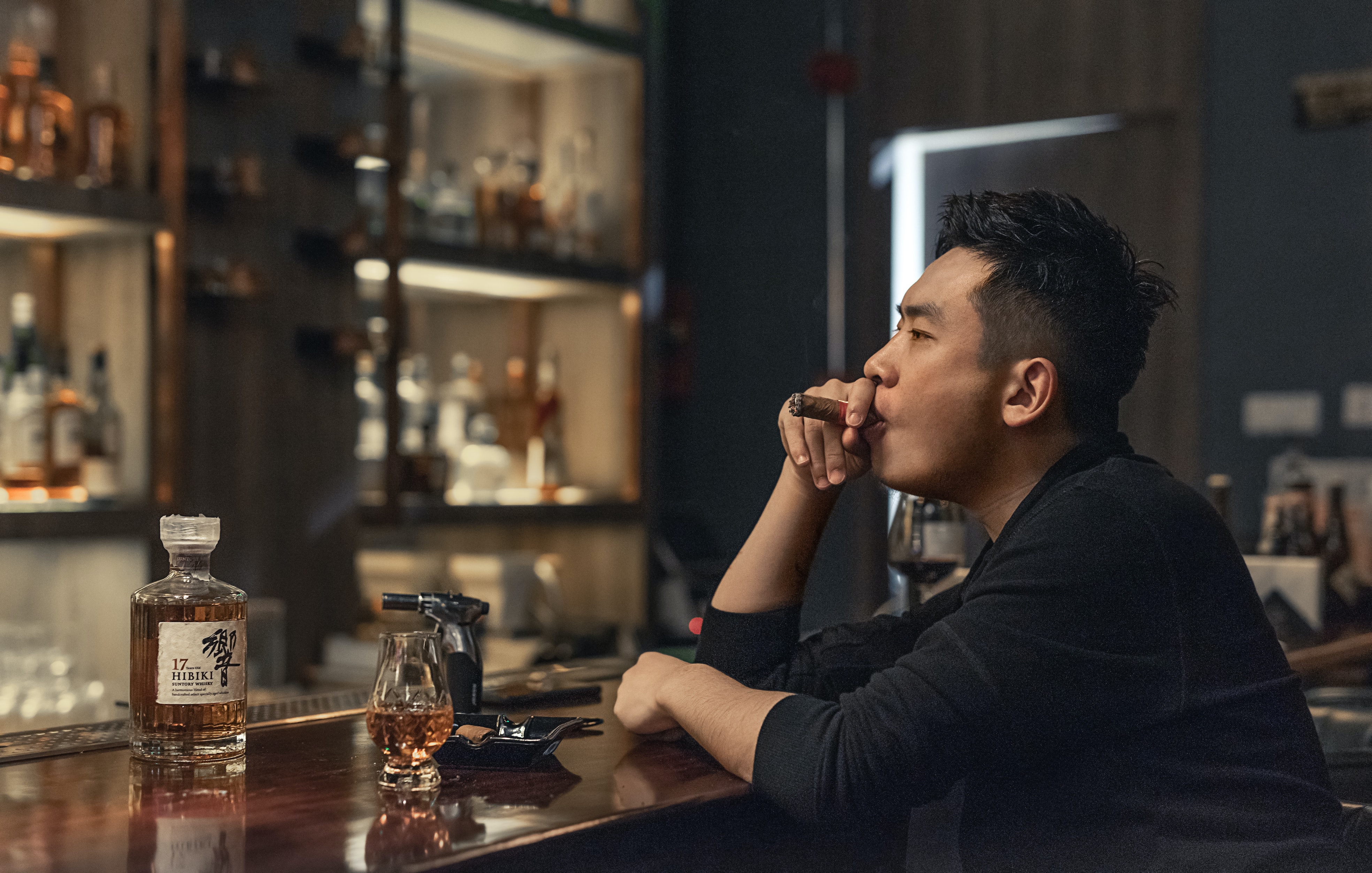 A man sat at a bar smoking a cigar and drinking whisky